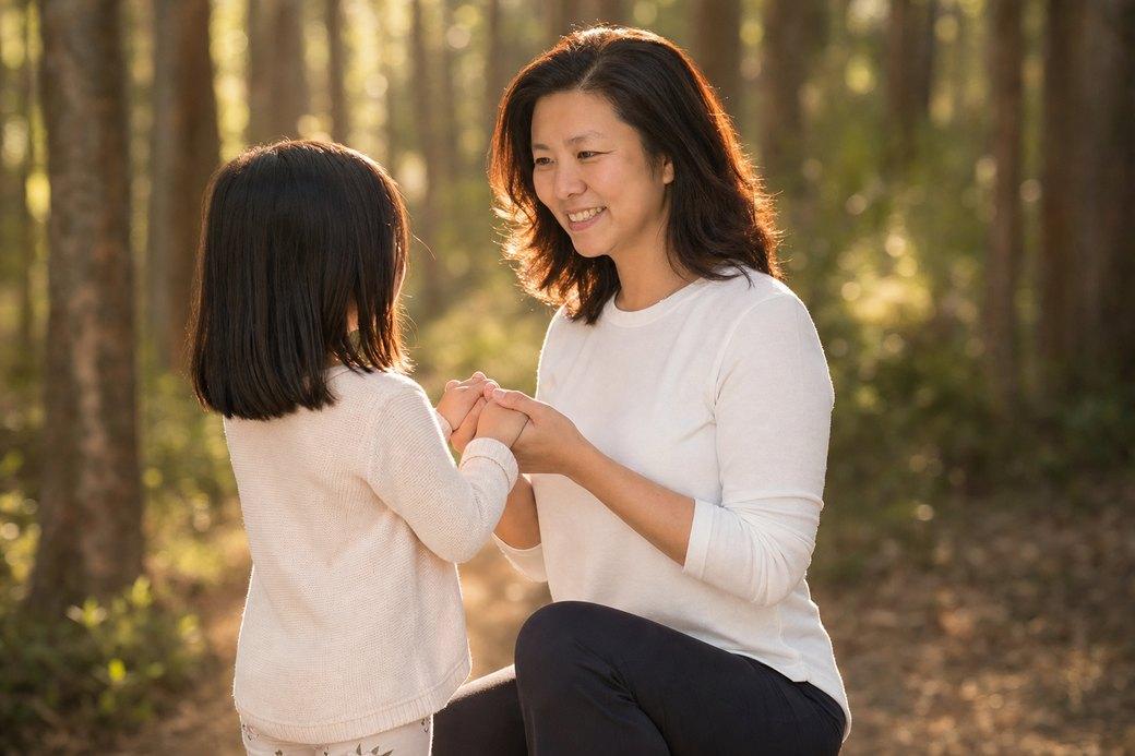 Dao, the founder of PlotJoys, kneeling in sunlit woodland holding both of her young daughter's hands
